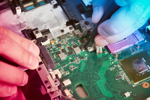 Man repairs computer. A service engineer in a shirt repairs a laptop, at a white Desk against a white wall. Computer motherboard background with blur neon multicolor glow. Hands close up.