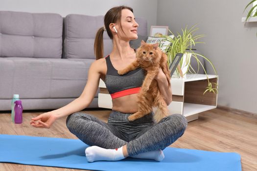Beautiful young woman in yoga posing with red cat indoor in flat.