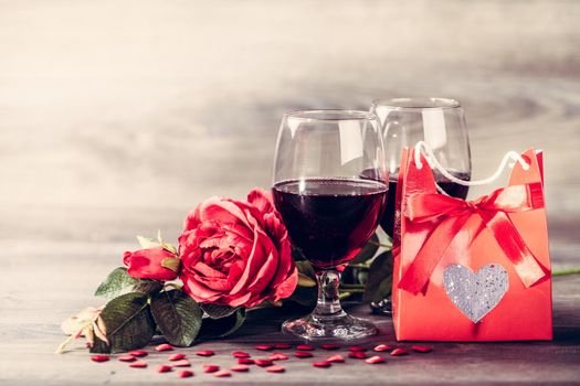 Two glasses of red wine, rose and gift with decoration by red hearts on wooden table. Valentines day celebration concept. Copy space.