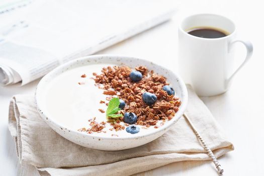 Breakfast with cup of coffee, newspaper, yogurt with chocolate granola, bilberry on white background, side view