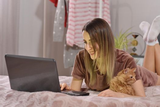 Young caucasian brunette businesswoman working at home using laptop while caressing pet red cat