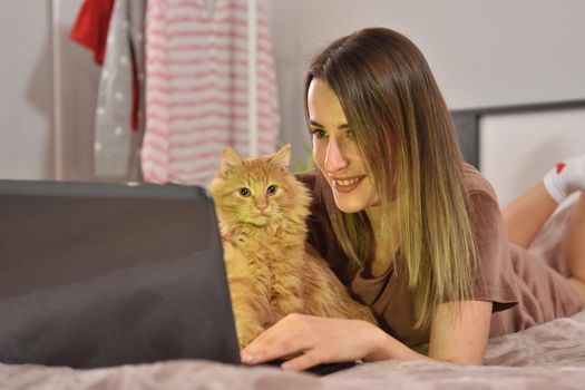 Young caucasian brunette businesswoman working at home using laptop while caressing pet red cat