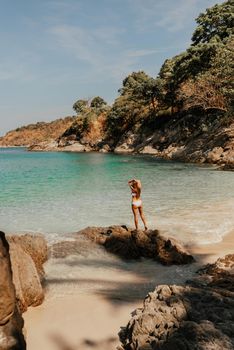 a young blonde European woman in a white bikini swimsuit on the beach stands backwards on the stone rocks on the ocean shore.