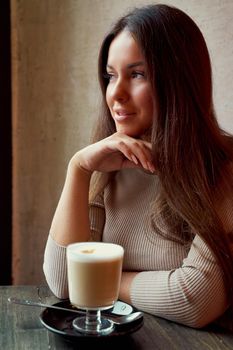 Beautiful pensive happy girl sitting in cafe in Christmas holidays, smiling and dreaming. Brunette woman with long hair drinks cappuccino coffee, latte and looks out window, vertical, dark backdrop