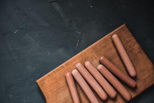 Boiled fried sausages sausages lie on a wooden kitchen board scratched against a dark concrete background.