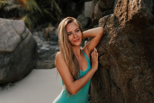Young blonde smiling European woman in bikini swimsuit on beach against background of stone rocks