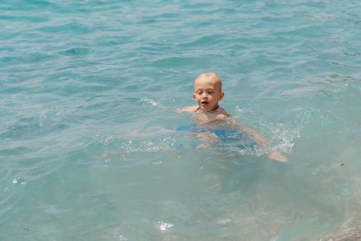 Child learning to swim in the open sea of tropical resort. Kids learn swimming. Exercise and training for young children. Little boy with colorful float board in sport club. Swimming baby or toddler. Happy child boy swims in sea in swimming circle with splash. Blue sky and water. Swimming training. Fun joy activities on vacation in the beach. Childhood moments lifestyle. Freedom careless. boy swim in the sea.