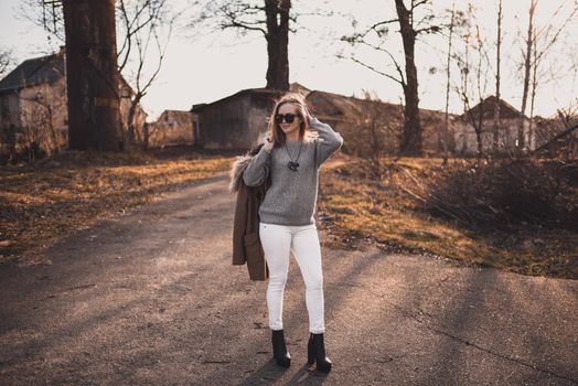 beautiful young blonde model girl. White pants. gray knitted sweater. black boots. black sunglasses. wooden pendant on the neck in the form of a horse. in a brown coat posing. On the Sunset. Portrait