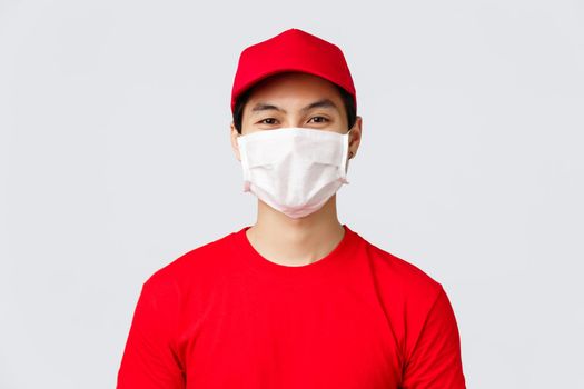 Close-up portrait of cheerful asian delivery guy in medical mask and red cap uniform, t-shirt, smiling with eyes at customer, taking order using safety measures to prevent covid-19 spread.