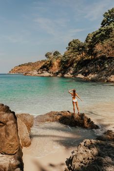 a young blonde European woman in a white bikini swimsuit on the beach stands backwards on the stone rocks on the ocean shore.