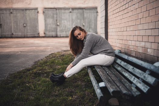 beautiful young blonde model girl sitting on a shabby bench. White pants. gray knitted sweater. black boots. wooden pendant on the neck in the form of a horse. on a background of gray wooden gates