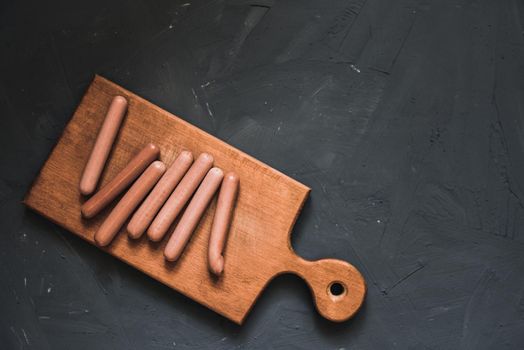 Boiled fried sausages sausages lie on a wooden kitchen board scratched against a dark concrete background.