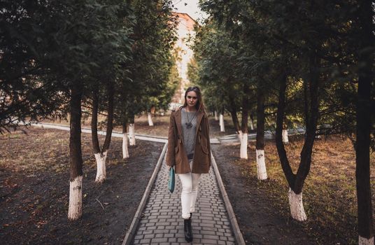 beautiful young blonde model girl. White pants. gray knitted sweater. black boots. . wooden pendant on the neck in the form of a horse. in a brown coat posing. On the Sunset. Portrait. near the trees