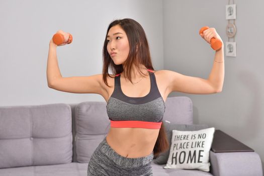 Young asian brunette woman in sportwear making home workout with two orange dumbbells. Soft focus