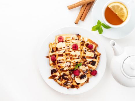 Belgian waffles with raspberries, banana, chocolate syrup. Breakfast with tea on white background, top view, copy space
