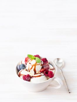 Balls of vanilla ice cream in mug with raspberry and blueberries, chocolate syrup topping. Side view, vertical, copy space, white bright background