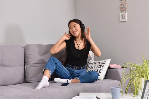 Excited beautiful asian teen listening music in headphones on her sofa at home. Wearing jeans and black tshirt