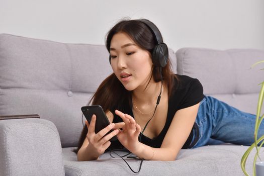 Excited beautiful asian teen listening music in headphones on her sofa at home. Wearing jeans and black tshirt