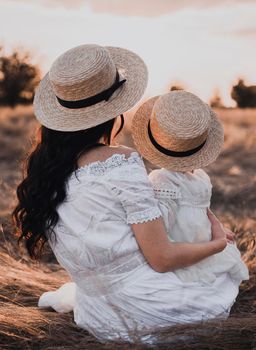 brunette mother and little daughter are sitting on the dry grass in white dresses. On the Sunset. in hats boater with his back.