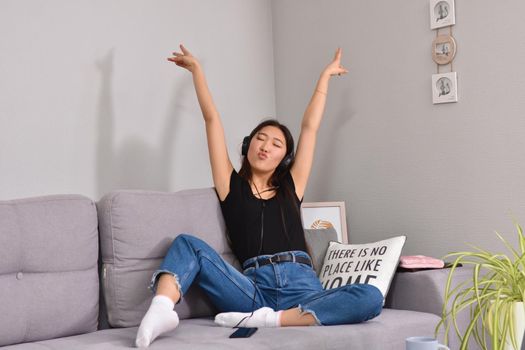 Excited beautiful asian teen listening music in headphones on her sofa at home. Wearing jeans and black tshirt