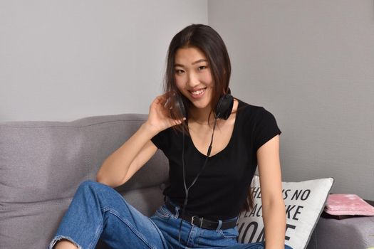 Excited beautiful asian teen listening music in headphones on her sofa at home. Wearing jeans and black tshirt