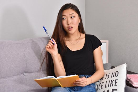 Attractive young asian female with long black hair relaxing on couch at home, writing down ideas for her project, using pen and copybook