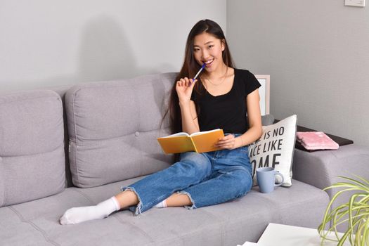 Attractive young asian female with long black hair relaxing on couch at home, writing down ideas for her project, using pen and copybook