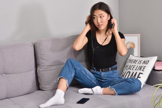 Excited beautiful asian teen listening music in headphones on her sofa at home. Wearing jeans and black tshirt