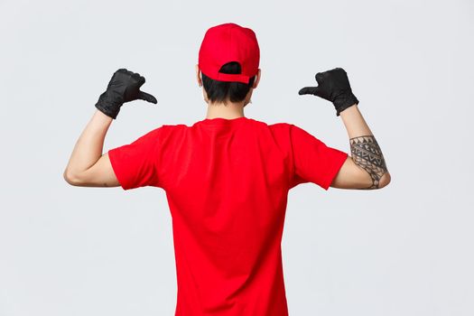 Back view of delivery man in red cap and t-shirt, wearing protective gloves during pandemic covid-19. Pointing at himself, show company logo uniform, ensure fast delivery, courier, shopping concept.