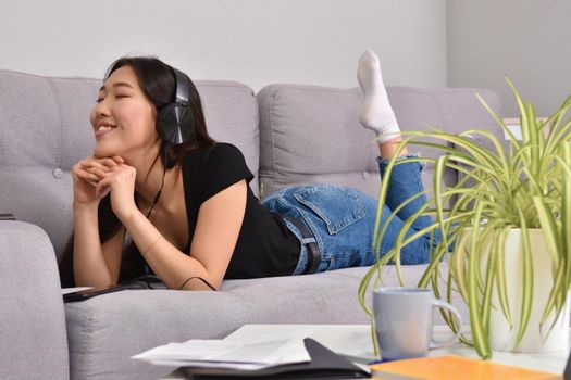 Excited beautiful asian teen listening music in headphones on her sofa at home. Wearing jeans and black tshirt