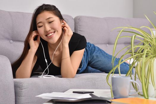 Excited beautiful asian teen listening music in headphones on her sofa at home. Wearing jeans and black tshirt