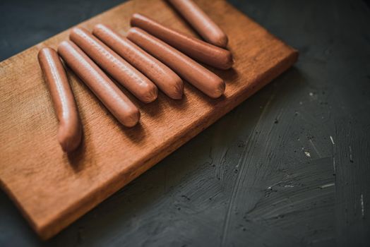 Boiled fried sausages sausages lie on a wooden kitchen board scratched against a dark concrete background.