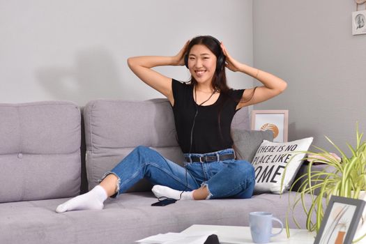 Excited beautiful asian teen listening music in headphones on her sofa at home. Wearing jeans and black tshirt