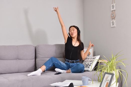 Excited beautiful asian teen listening music in headphones on her sofa at home. Wearing jeans and black tshirt