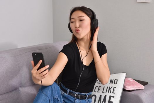 Excited beautiful asian teen listening music in headphones on her sofa at home. Wearing jeans and black tshirt