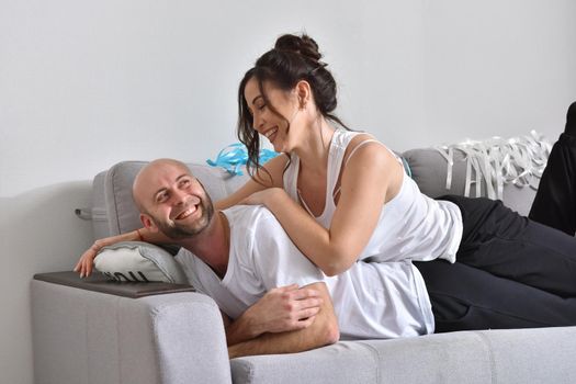 Photo of cheerful romantic caucasian family couple smiling and hugging while sitting on sofa at home. Man and woman in love