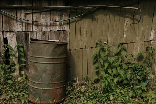 Old wooden shabby frail little fishing house. Under the roof of the house is attached a fishing rod for fishing. Near the house there is a huge Green barrel for water. Old iron metal around. Green big