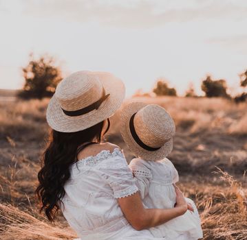 brunette mother and little daughter are sitting on the dry grass in white dresses. On the Sunset. in hats boater with his back.