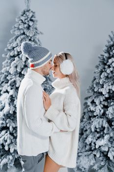 Couple kissing near christmass trees. Winter holidays. Love story of young couple weared white pullovers. Happy man and young woman hug each other.