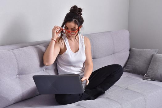 Beautiful brunette woman in red glasses on sofa using laptop