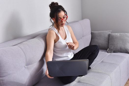 Beautiful brunette woman in red glasses on sofa using laptop