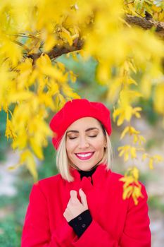 Optimistic young stylish female with blond hair and makeup, wearing red beret and coat, while standing under bright yellow tree in park on sunny autumn day