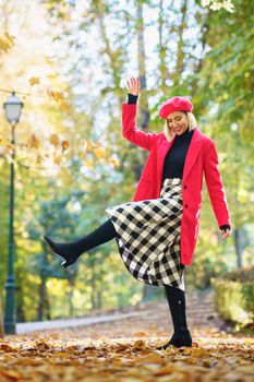 Full body of positive female in red coat and beret raising leg while standing on pathway with fallen leaves in park