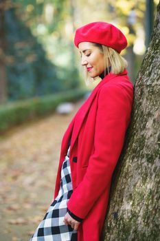 Side view of female wearing red beret and coat with checkered skirt standing near tree trunk in autumn park and looking down