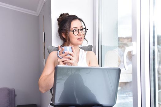 Home quarantine, protection from coronavirus. Caucasian brunette woman sit on window with computer