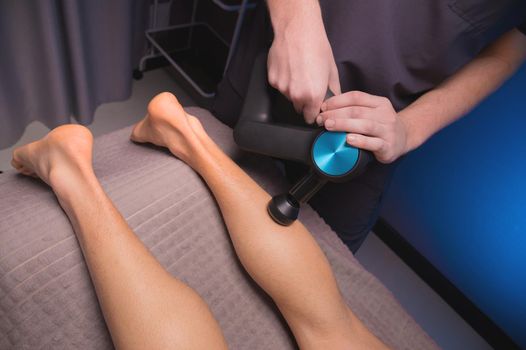 A professional masseur makes a massage of the calf muscle with a massage gun to a man athlete in a massage room with low light.