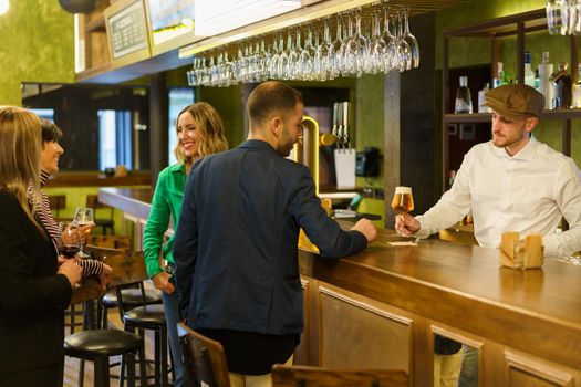 Man ordering beer from barkeeper while standing near happy talking women on weekend day in pub