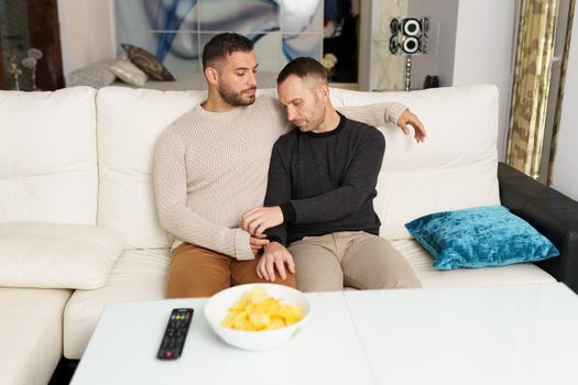 LGBT couple of handsome homosexual men hugging on couch in living room and chilling together at weekend at home