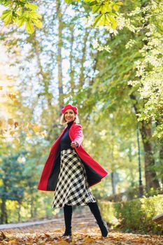 Full body of active female in red coat and beret looking at camera while standing on pathway on autumn day