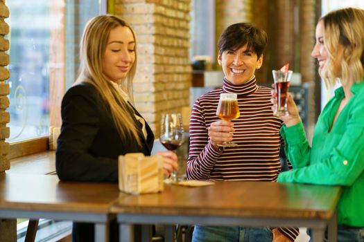 Merry adult female in casual clothes drinking beer and looking at camera with smile while spending time with girlfriends at table near window in pub